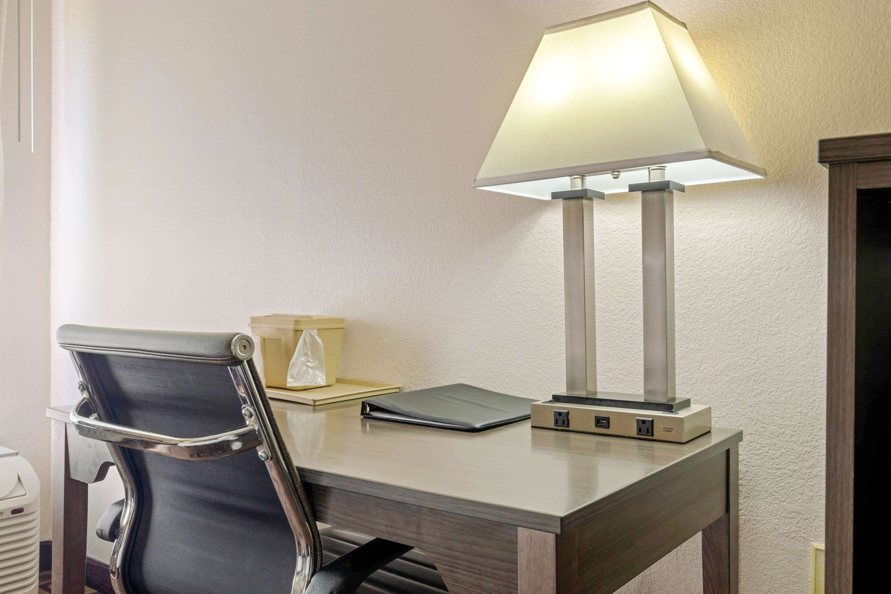 Escritorio de hotel con una silla, una lámpara, un adaptador de corriente y una caja pequeña.