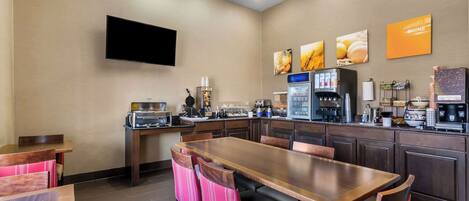 A hotel dining area with a long wooden table, multiple chairs, a flat-screen TV mounted on the wall, and a coffee machine.