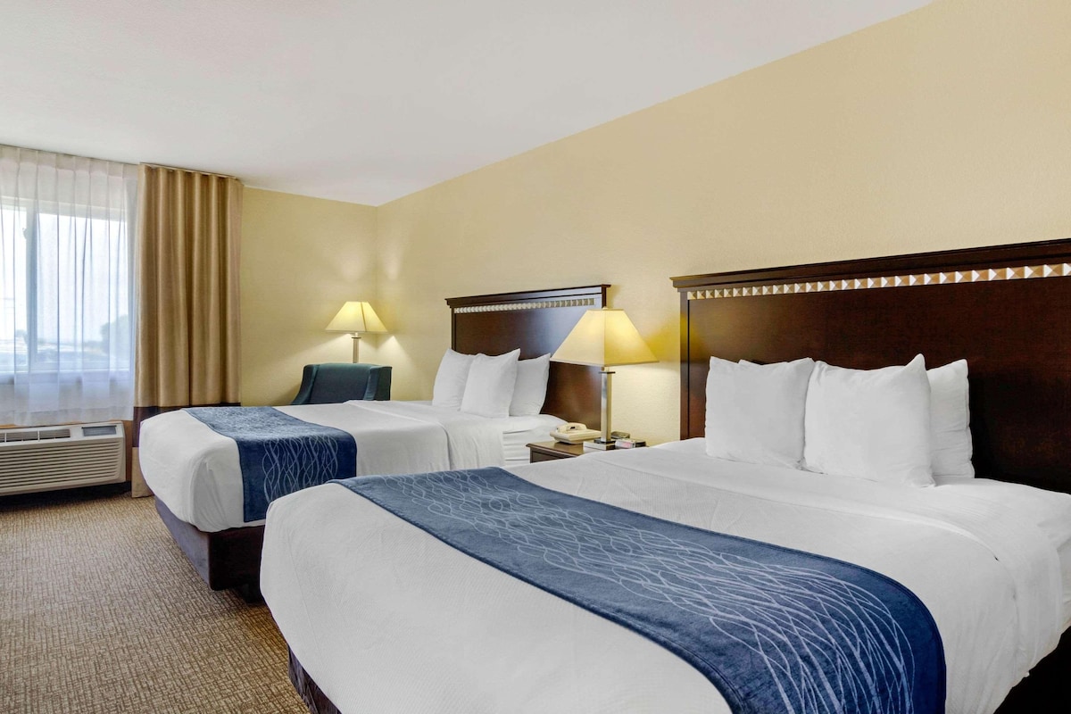A hotel room with two beds, a large window with curtains, a desk with a lamp, and an air conditioning unit.