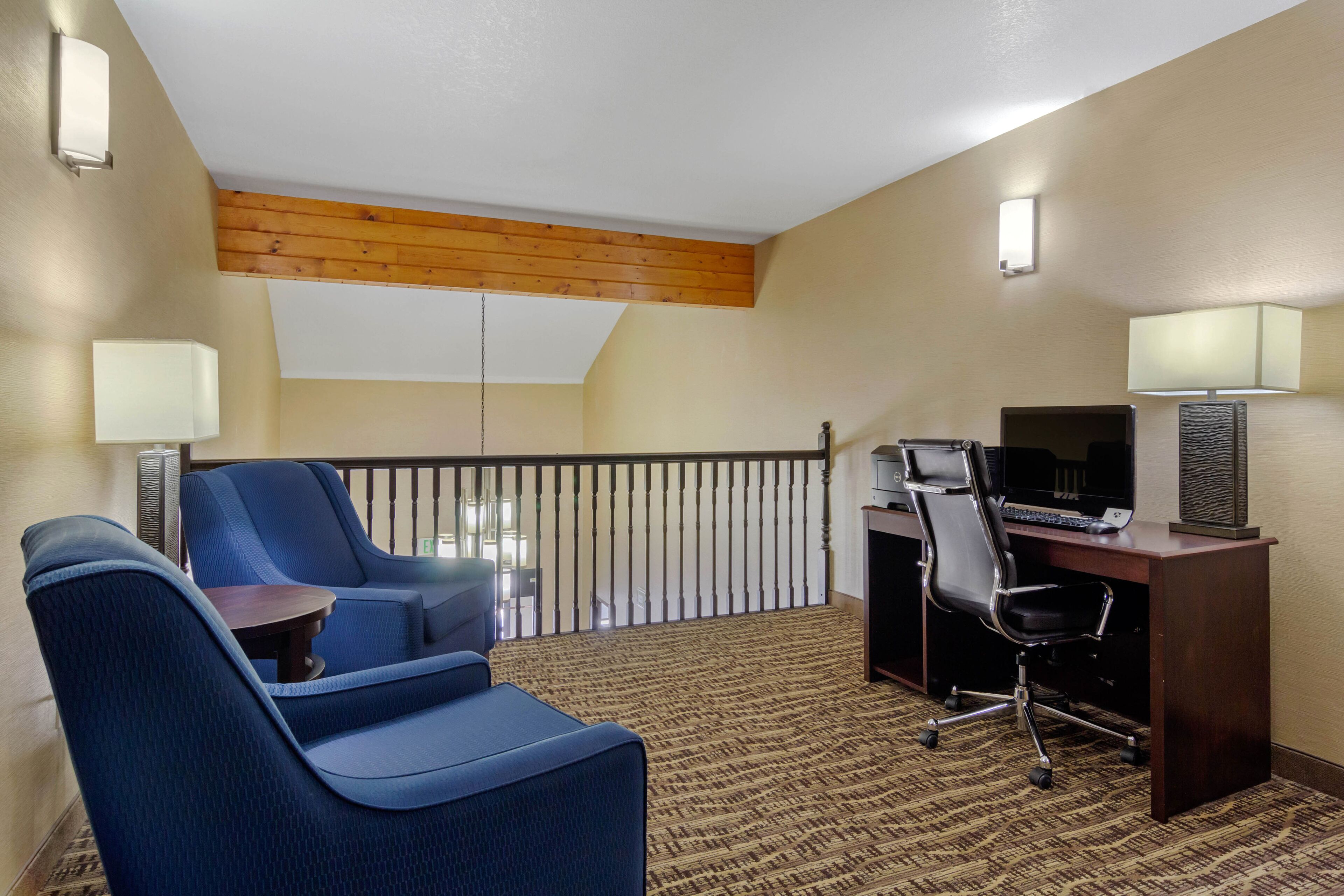Une chambre d’hôtel avec un balcon, un bureau avec un ordinateur, deux fauteuils bleus et une moquette à motifs.