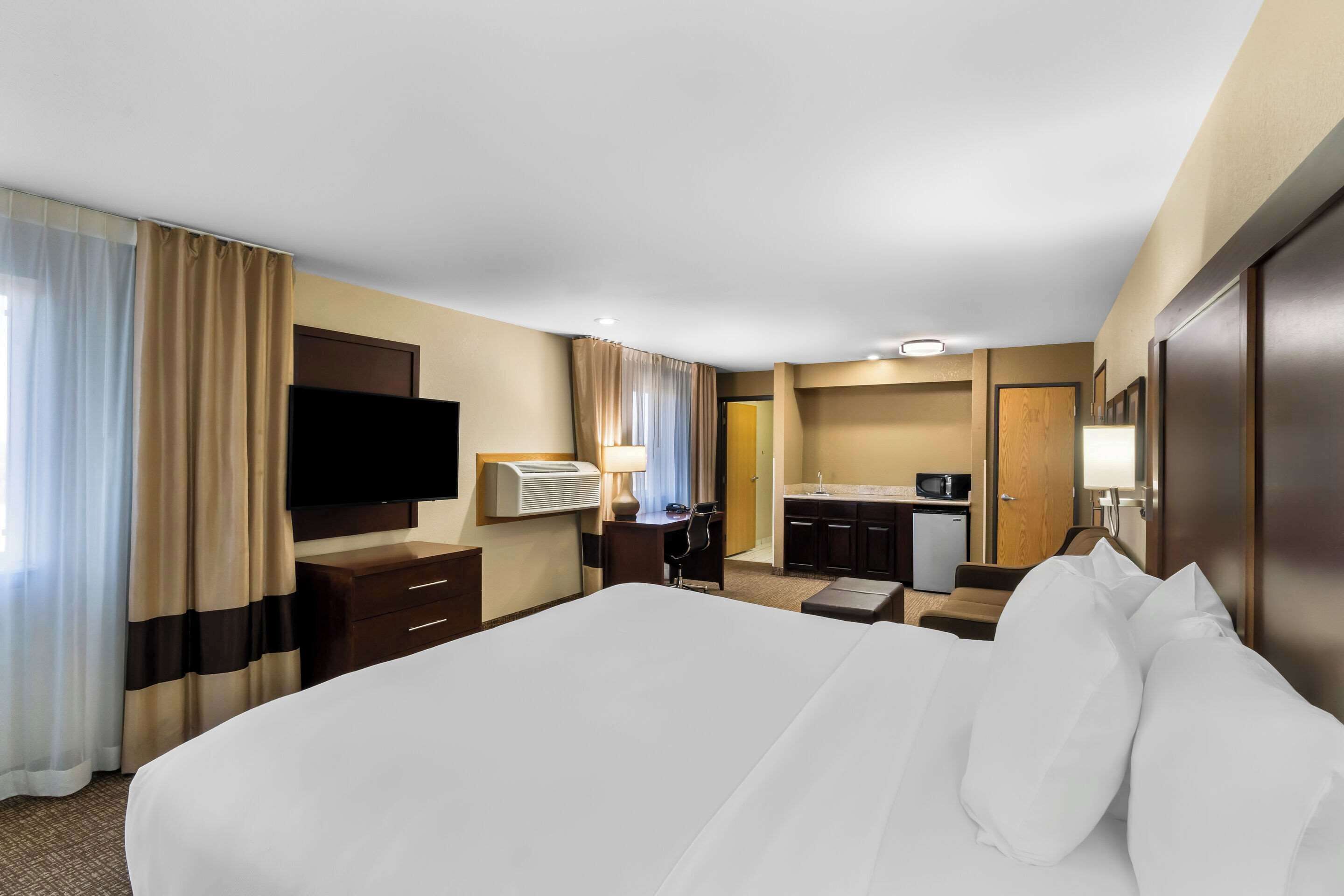 A hotel room with a large bed, a desk with a chair, a flat-screen TV, a minibar, and a bathroom visible through an open door.