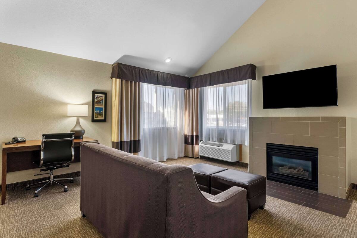 A hotel room with a sofa, a desk with a lamp, a flat-screen TV, a fireplace, and large windows with curtains.