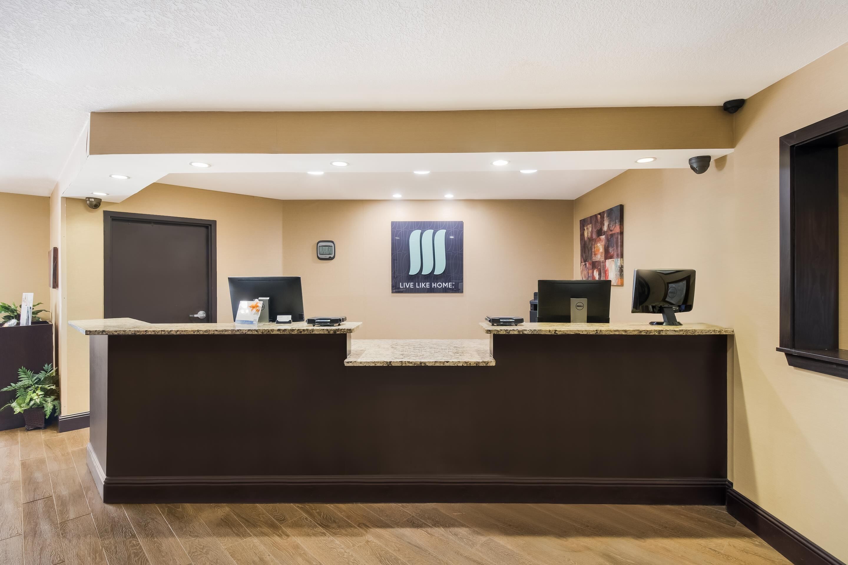 A reception desk with a "Live Like Home" sign, a security camera, and a framed artwork on the wall.