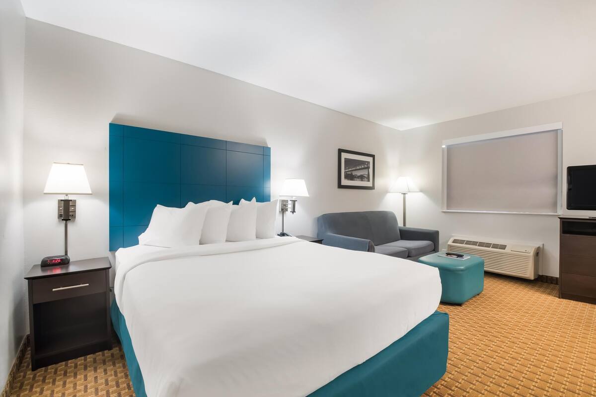 A hotel room with a large bed, two bedside tables with lamps, a gray armchair, a small ottoman, a TV, and a framed picture on the wall.