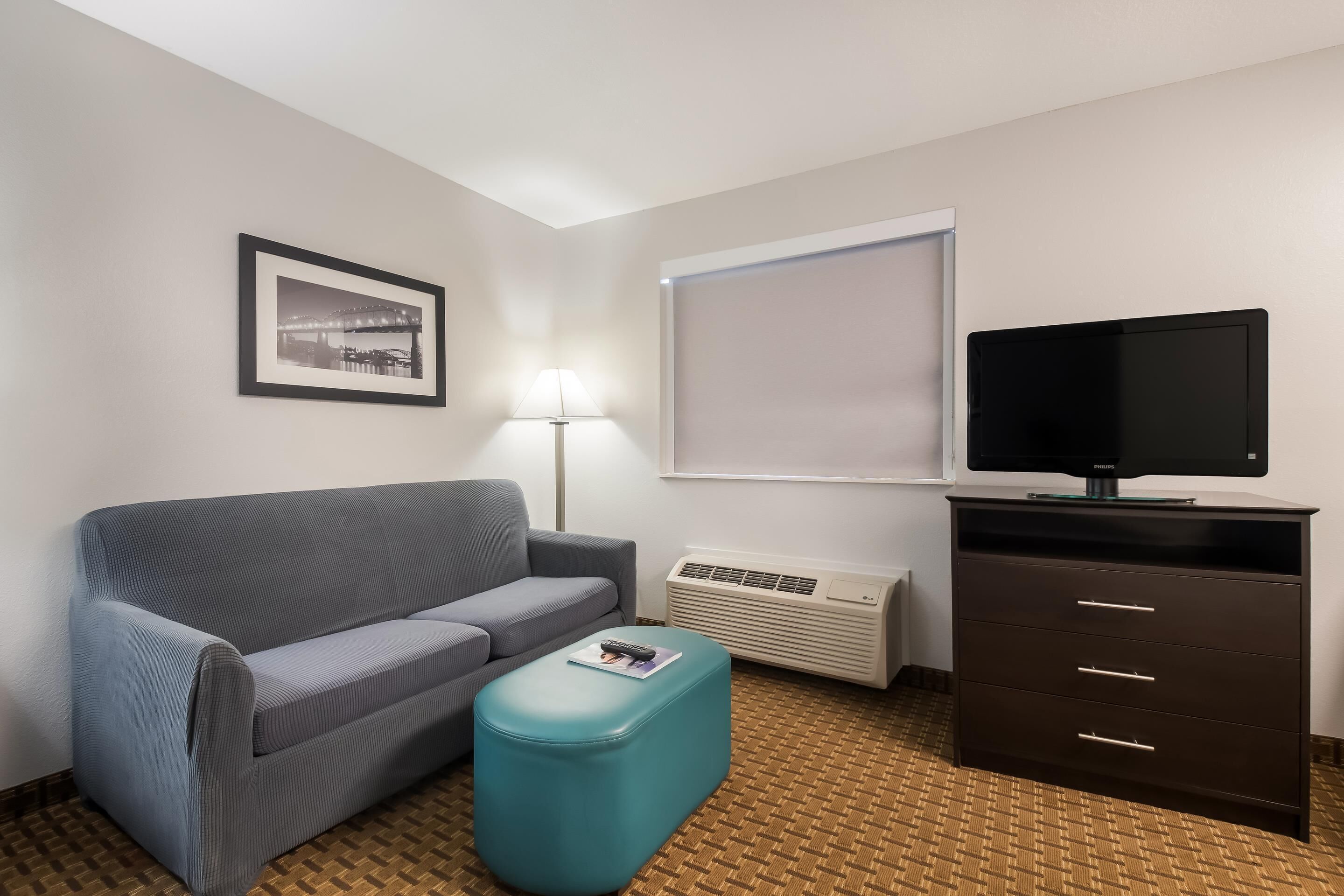 A hotel room with a grey sofa, a teal ottoman, a flat-screen TV on a dark cabinet, a framed picture on the wall, and a window with blinds.