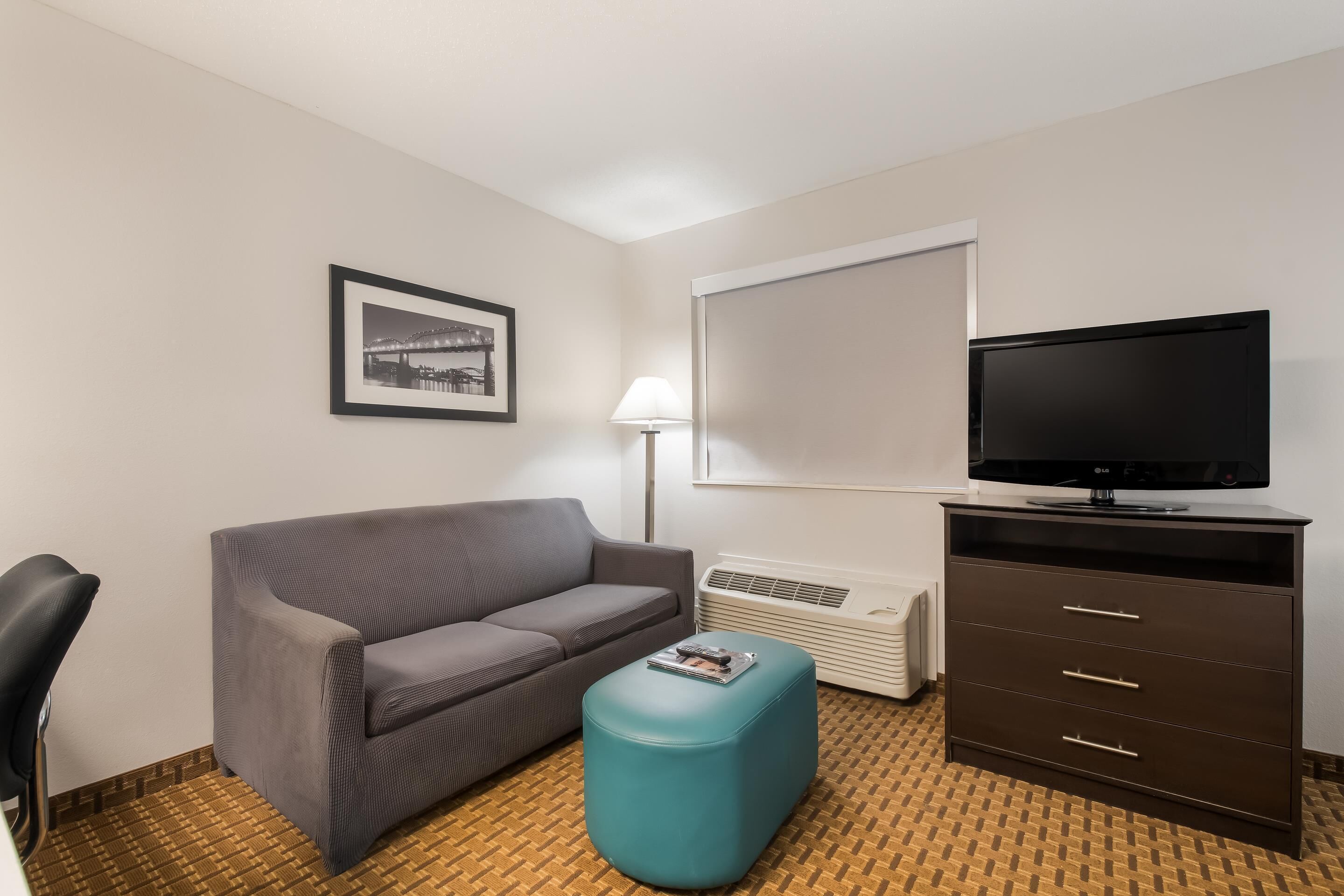 Une chambre d’hôtel avec un canapé gris, un pouf turquoise, un téléviseur posé sur un meuble, un tableau encadré au mur et un sol à motifs.