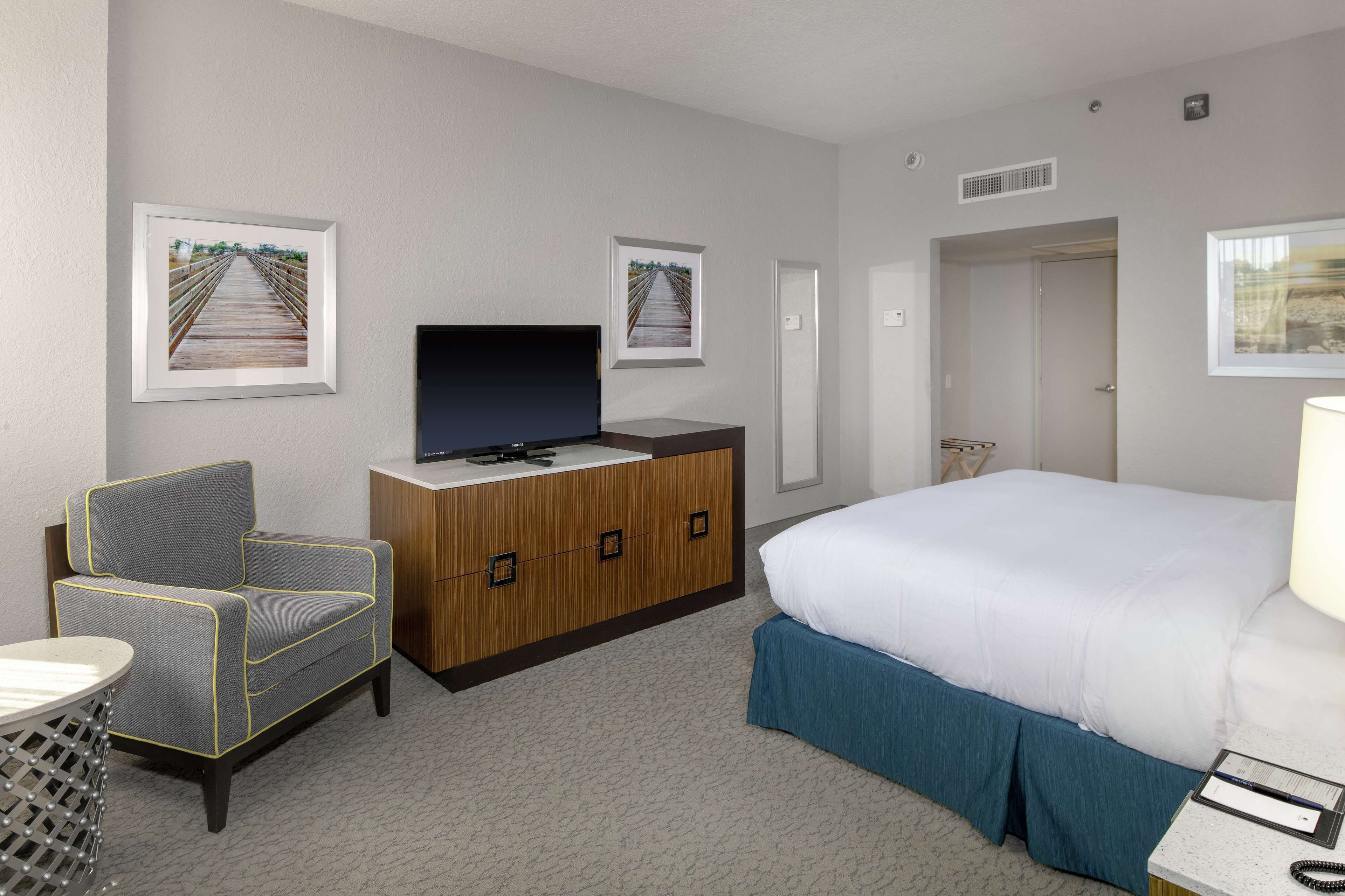 Una habitación de hotel con una cama, un televisor sobre un mueble de madera, un sillón gris y dos cuadros en la pared.