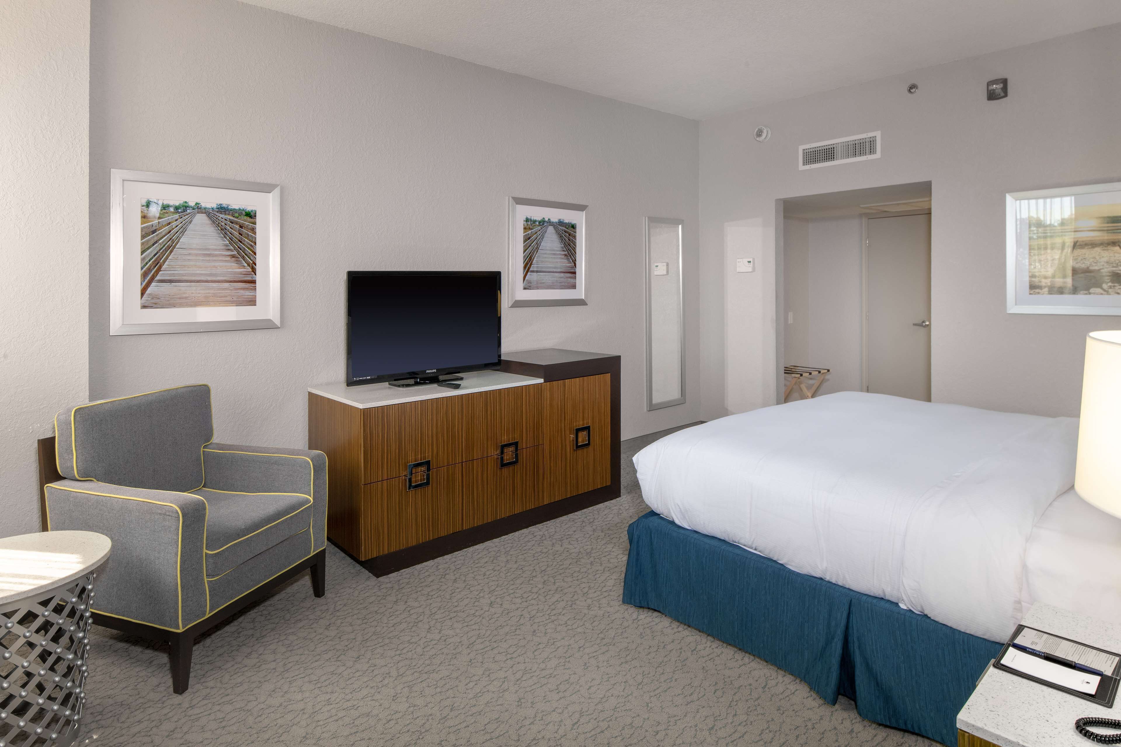 A hotel room with a bed, a TV on a wooden cabinet, a grey armchair, and two framed pictures on the wall.