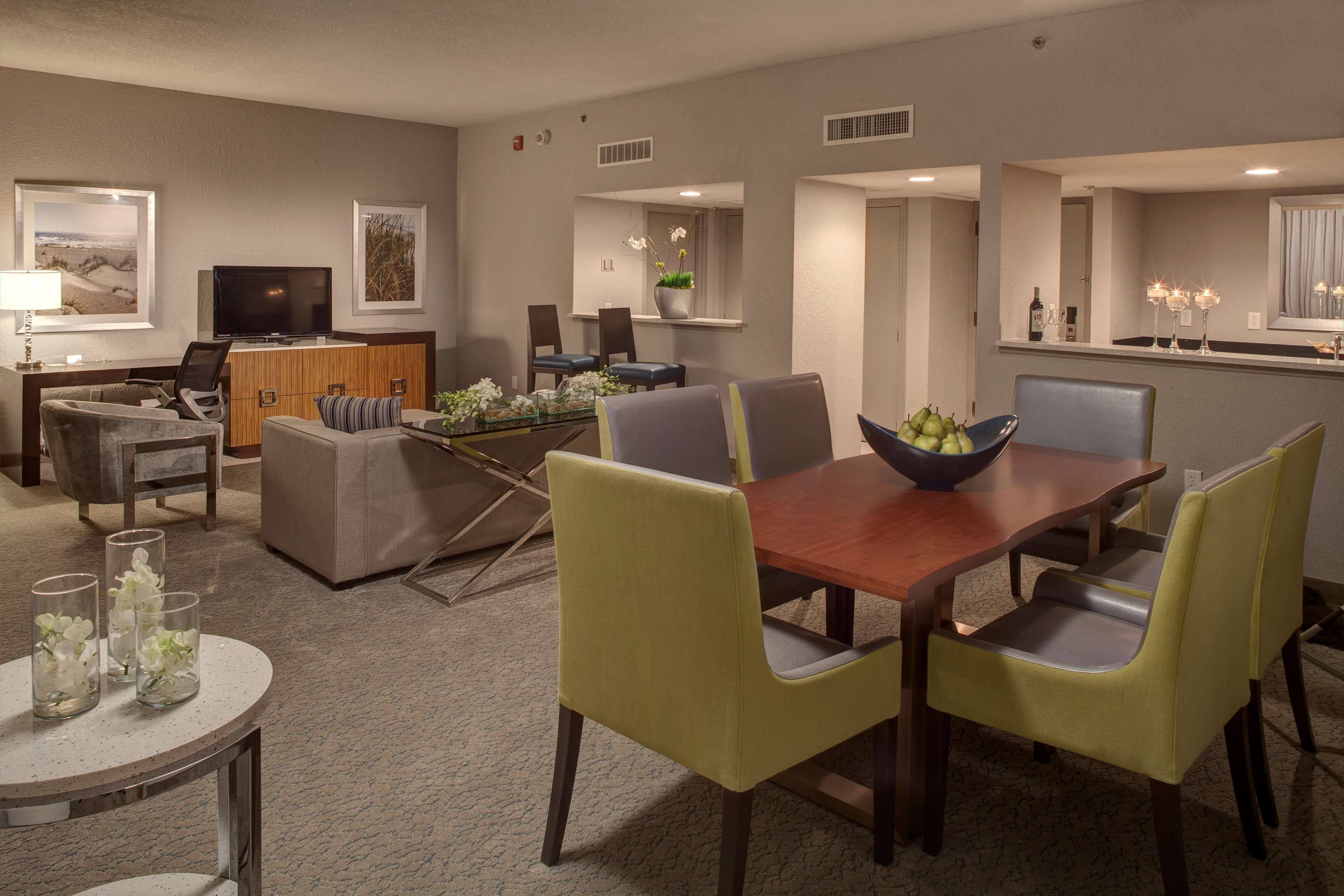 A modern hotel room with a dining area, a TV, a bar, and a view of the beach.