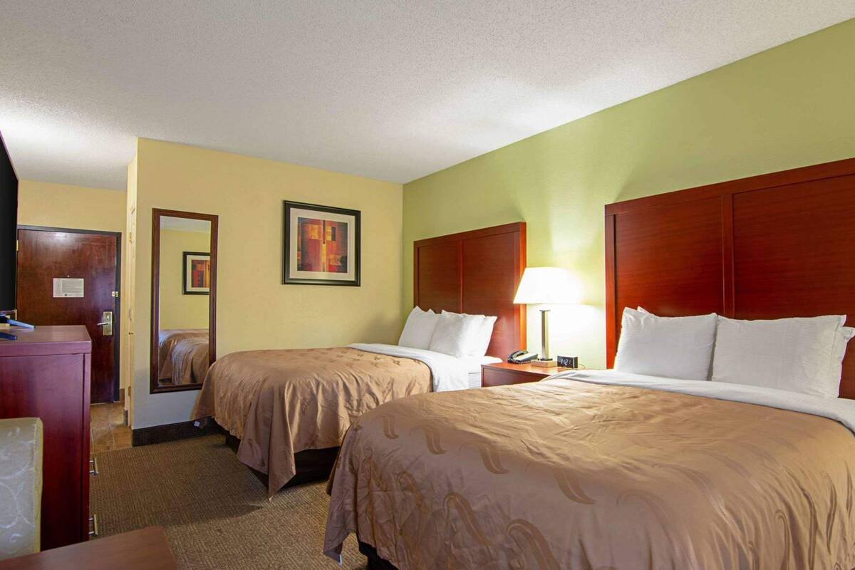 A hotel room with two beds, a desk, a mirror, and a lamp.
