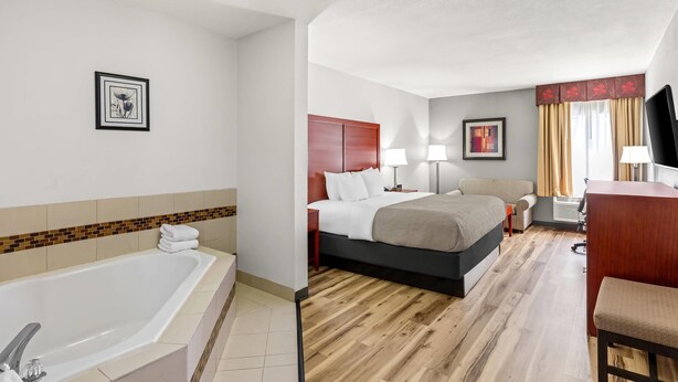 A hotel room with a large bed, a desk, a chair, a desk lamp, a TV, a bathroom with a bathtub, and a picture on the wall.