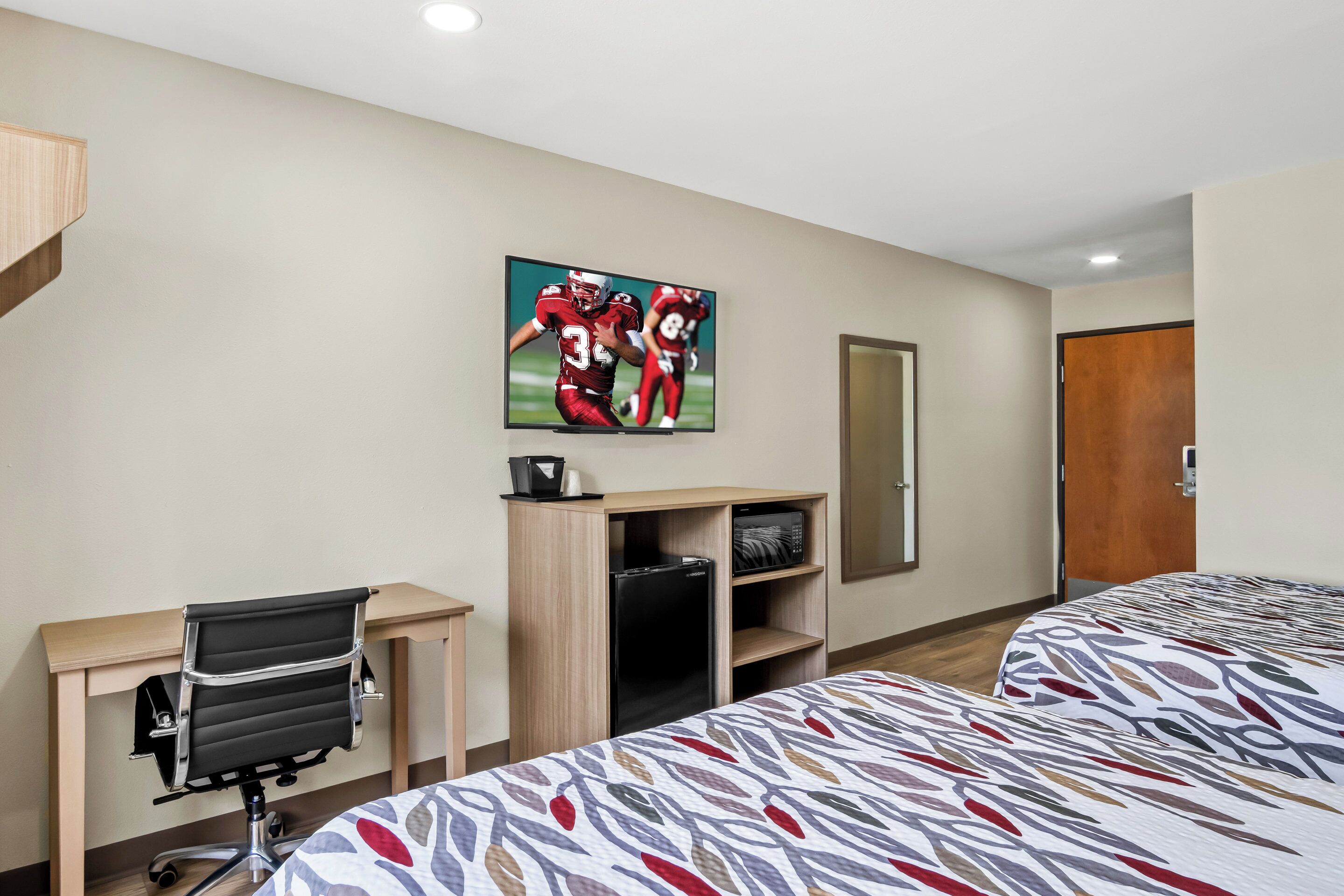 A hotel room with a bed, a desk with a chair, a refrigerator, a microwave, and a television on the wall.