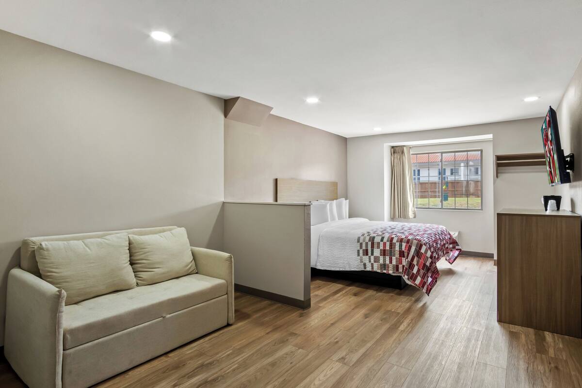 Un dormitorio moderno con piso de madera, una cama con colcha, un sofá, un escritorio y una ventana con cortinas.