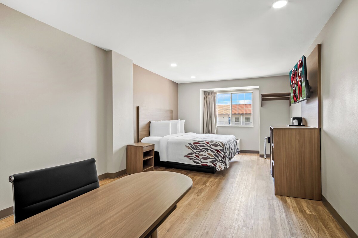 Ein modernes Hotelzimmer mit Holzboden, einem Bett mit gemustertem Überwurf, einem Schreibtisch, einem Stuhl, einer Heizung, einem Kleiderschrank und einem Fenster mit Vorhängen.