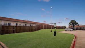 A fenced yard with a grassy area, a wooden fence, and a building with a red roof. - Red Roof Inn & Suites Euless - DFW Airport Southwest (Euless)