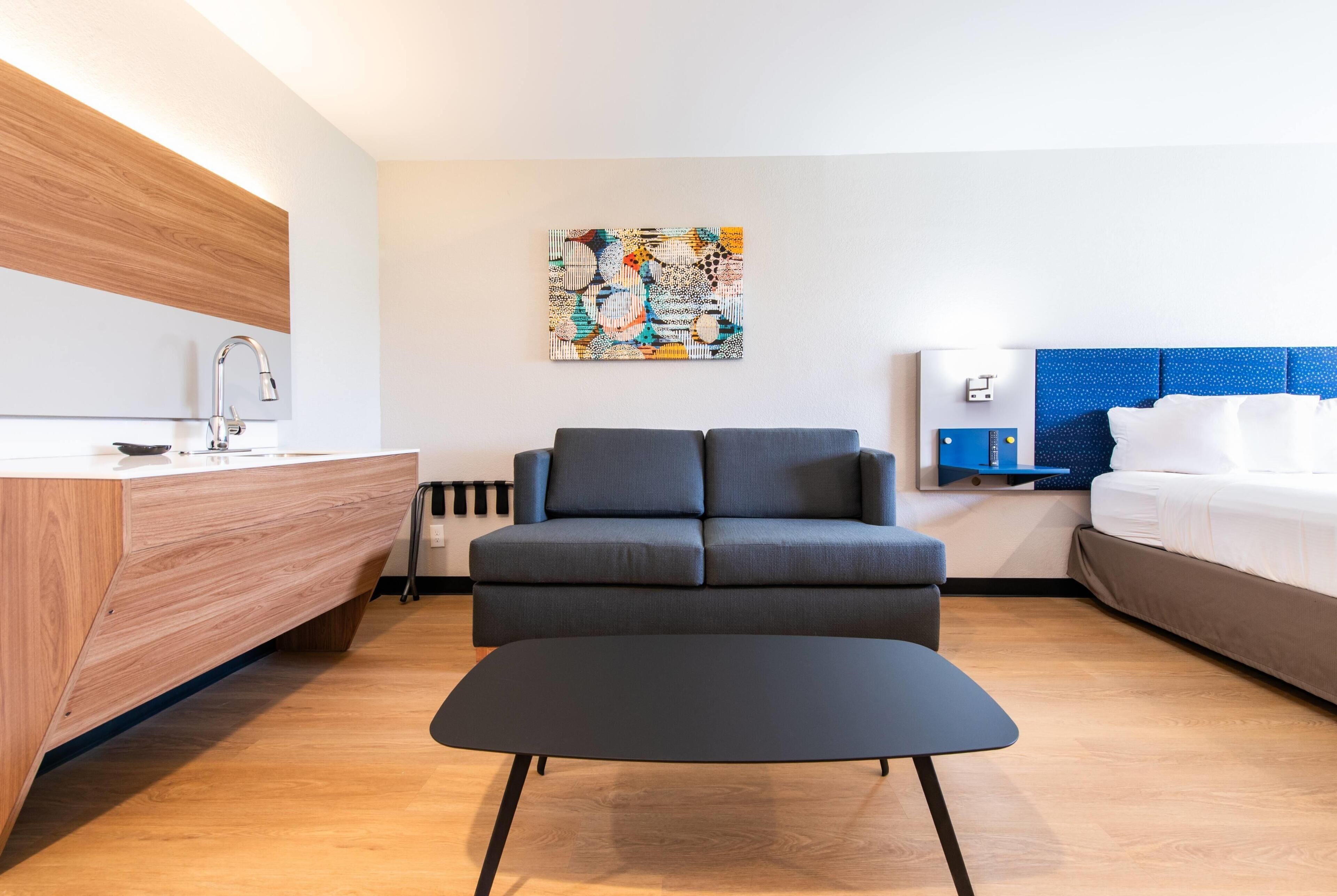 Una moderna camera d'hotel con una toeletta in legno, un divano grigio, un poggiapiedi nero, una testiera azzurra con pois e un quadro astratto colorato appeso al muro.