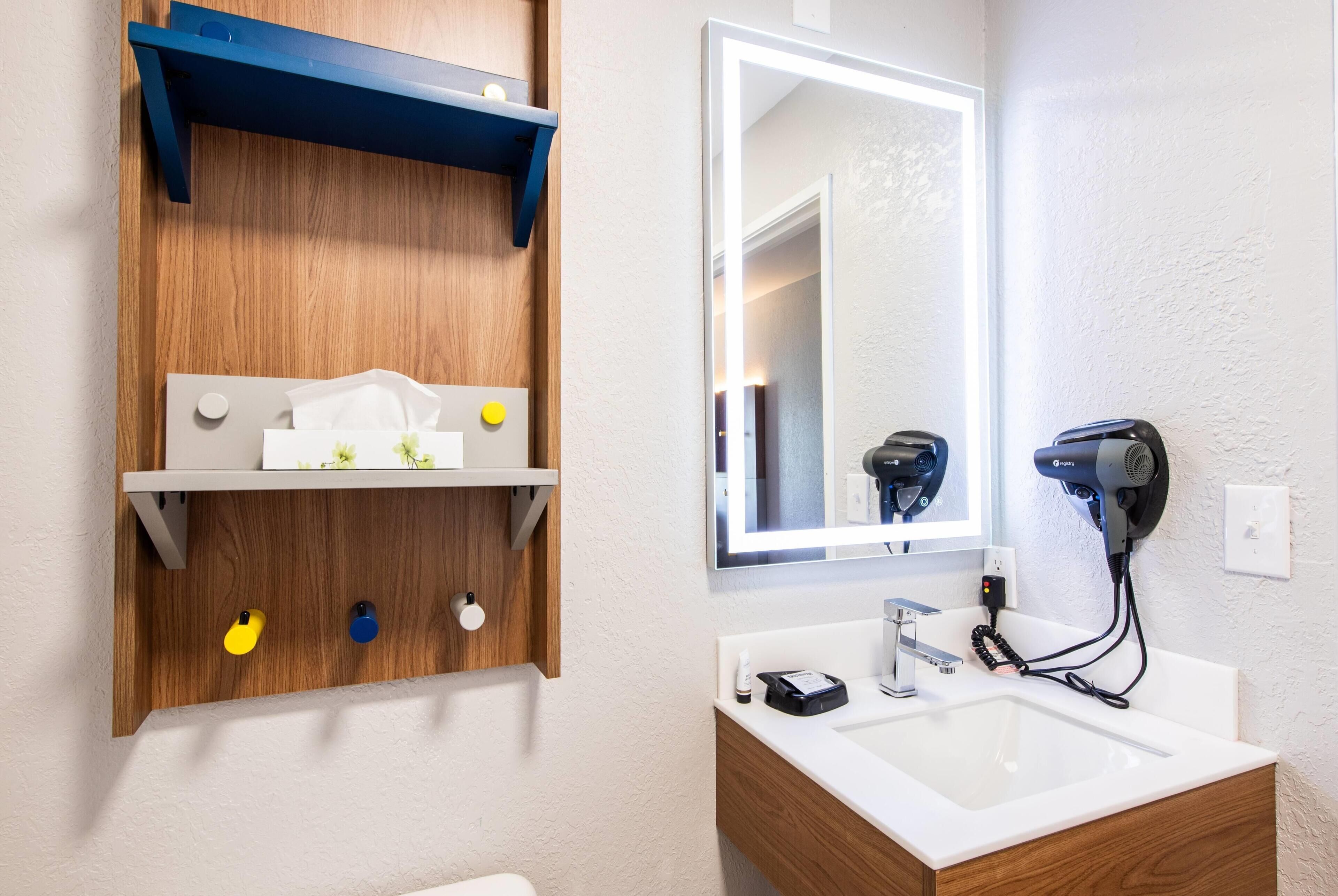 A bathroom with a white sink, a mirror, a hairdryer, and a wooden shelf with toiletries.