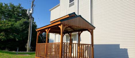 A wooden gazebo with a covered porch, adjacent to a white building with a window and a door.