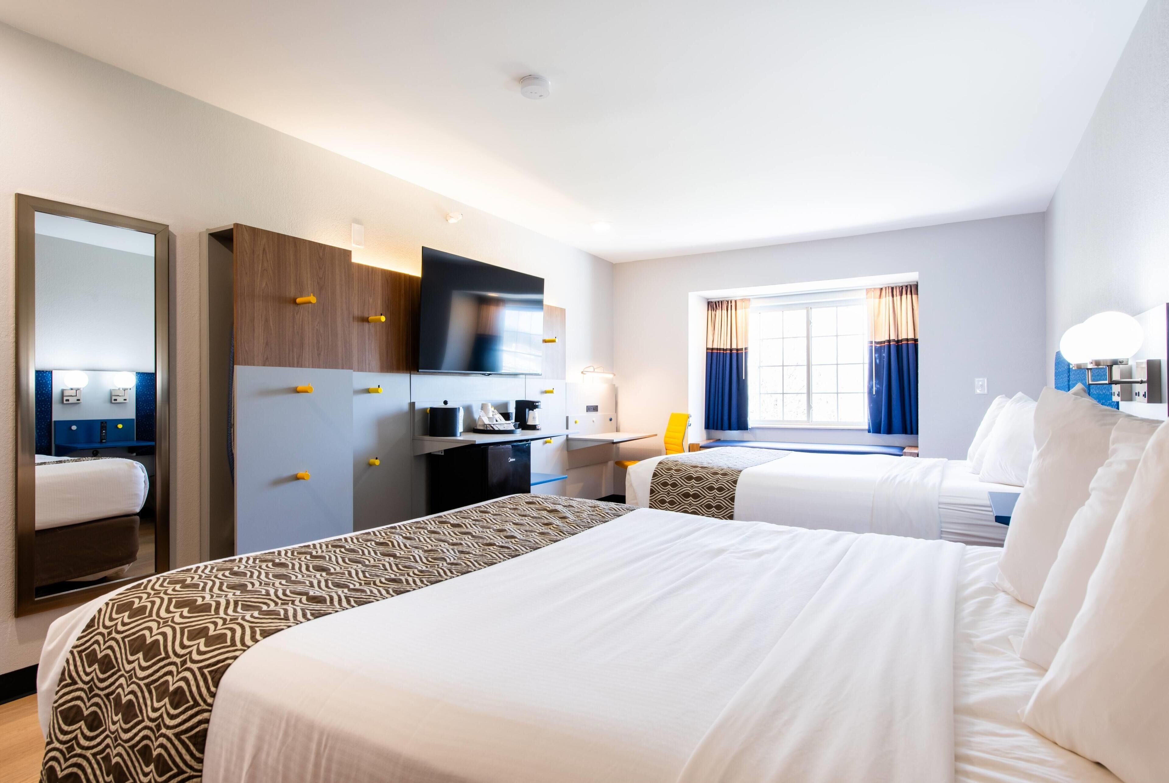 A hotel room with two beds, a desk, a TV, and a window with curtains.