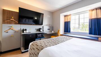 A hotel room with a bed, a TV mounted on the wall, a desk with a microwave, and a window with curtains.