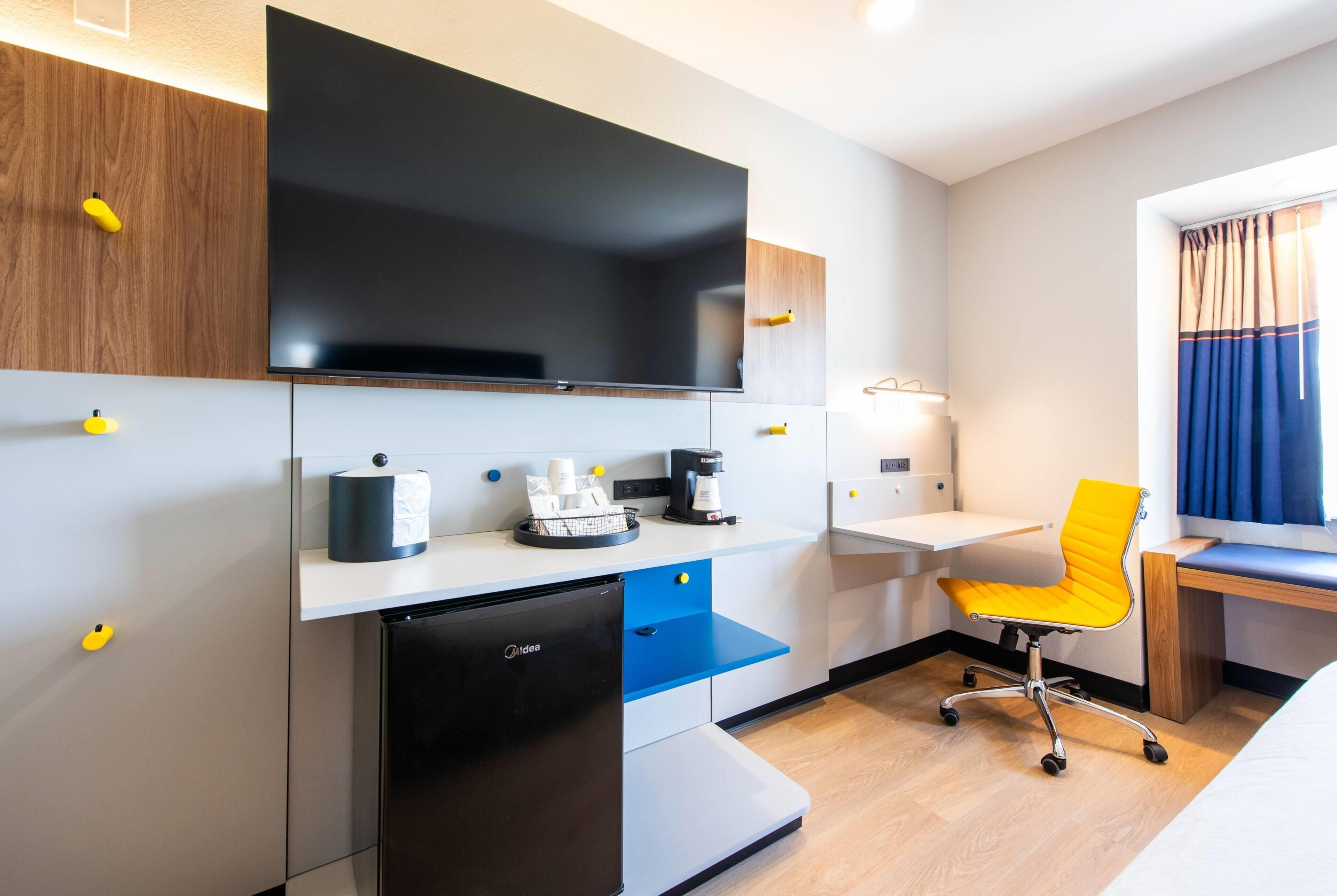 Una habitación de hotel compacta con kitchenette, televisor de pantalla plana, escritorio, silla y cama.