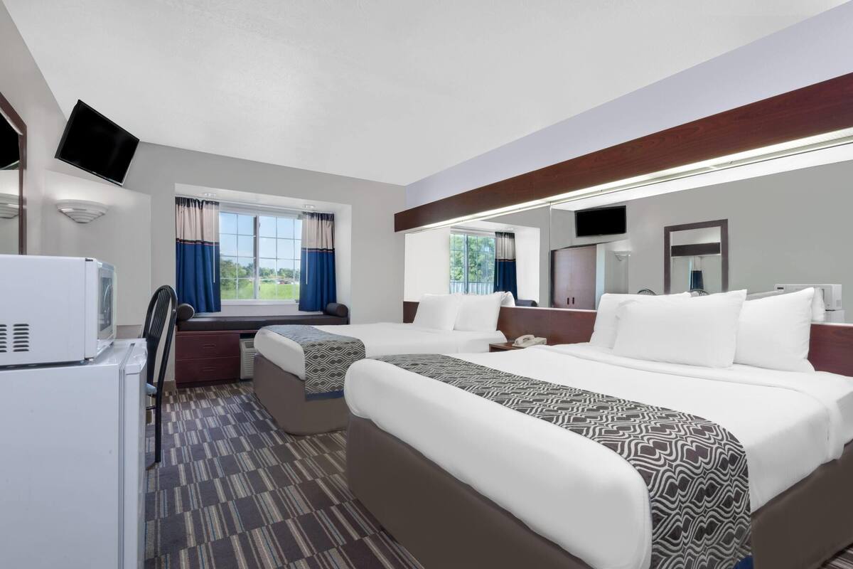A hotel room with two beds, a TV, a refrigerator, and a window with curtains.