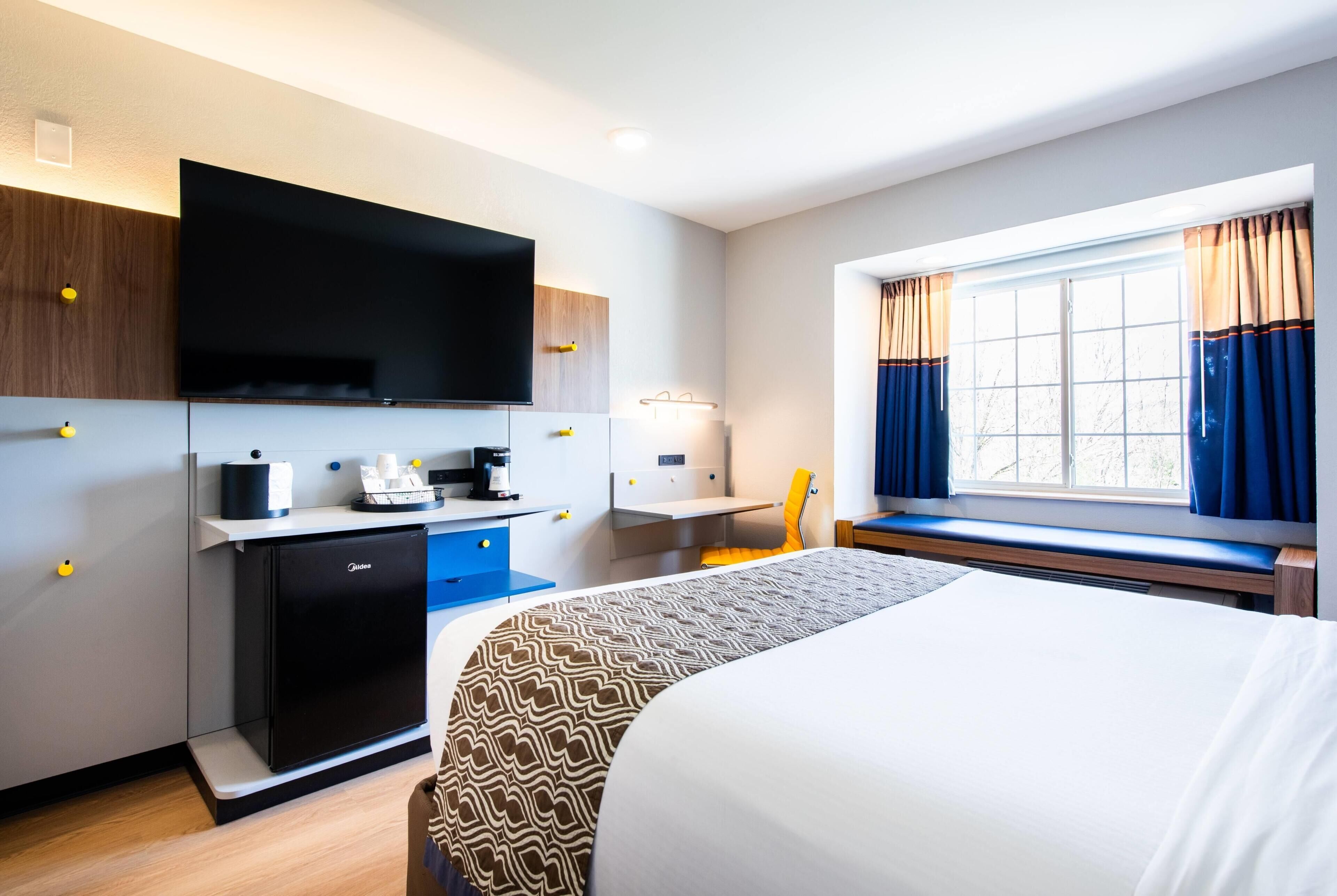 Une chambre d’hôtel avec un lit, un bureau avec un ordinateur, une télévision et une fenêtre avec des rideaux.