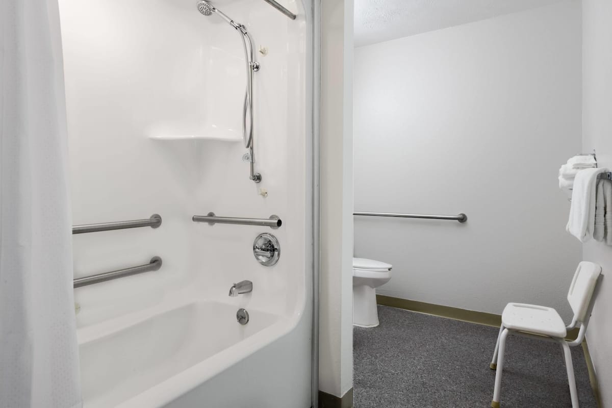 A bathroom with a white bathtub, a white toilet, a white chair, and grab bars.
