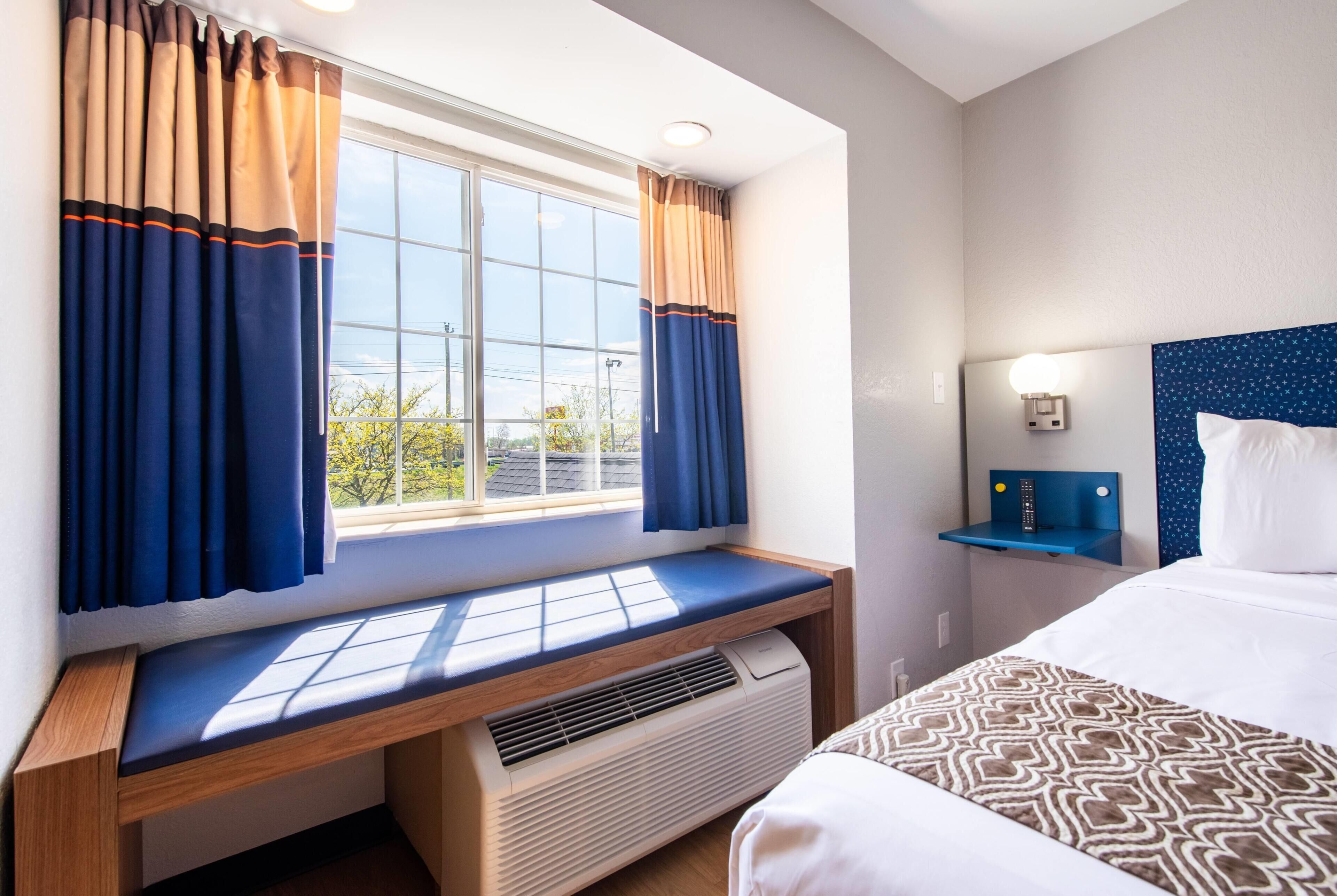 A hotel room with a bed, a window with curtains, a bedside table, and a wall-mounted lamp.