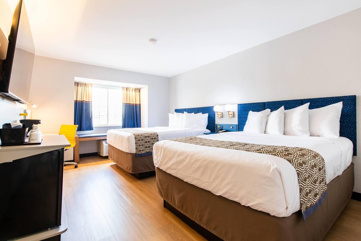 A hotel room with two beds, a desk, a chair, a window with curtains, and a flat-screen TV.