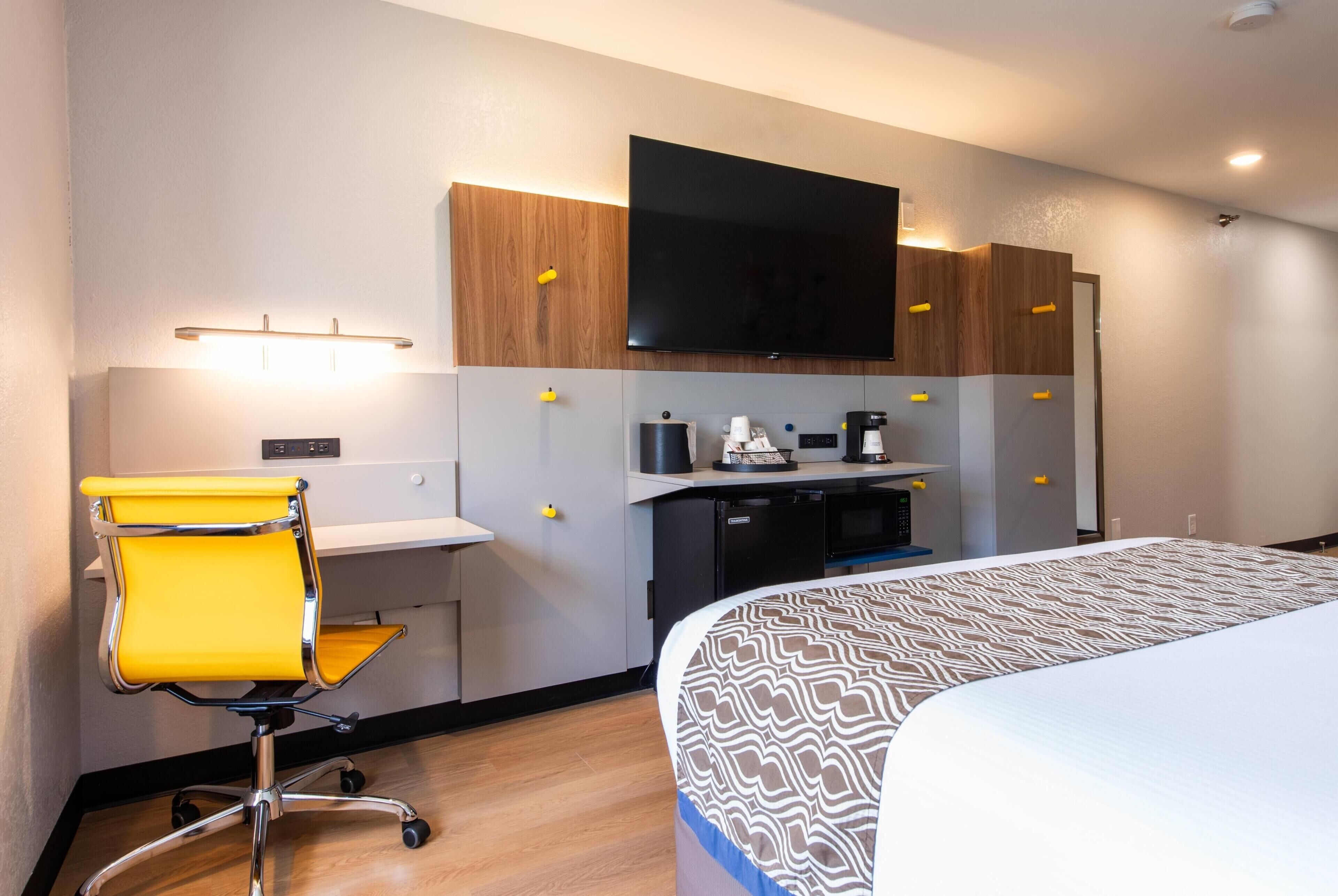 A hotel room with a bed, a desk with a chair, a flat-screen TV, a coffee machine, and a small kitchenette.