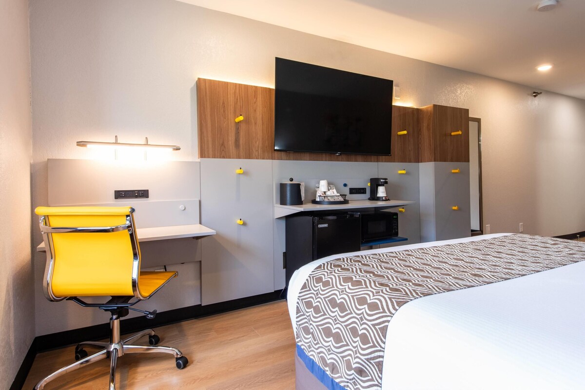 A hotel room with a bed, a desk with a chair, a flat-screen TV, a coffee machine, and a small kitchenette.