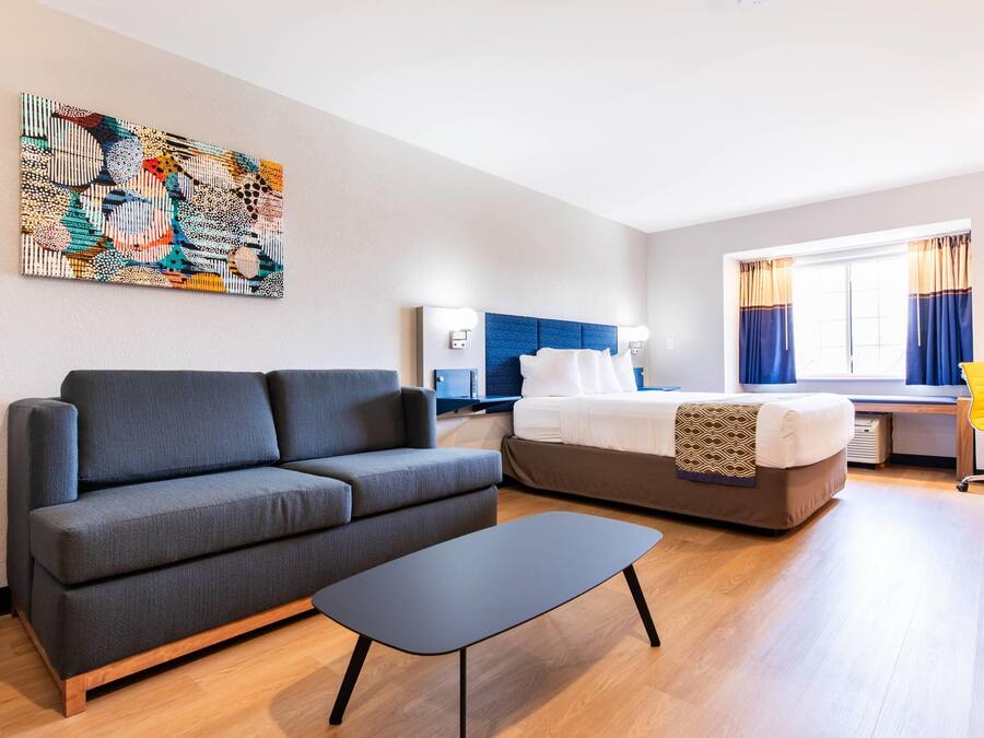 A modern hotel room with a sofa, bed, desk, and artwork on the wall.