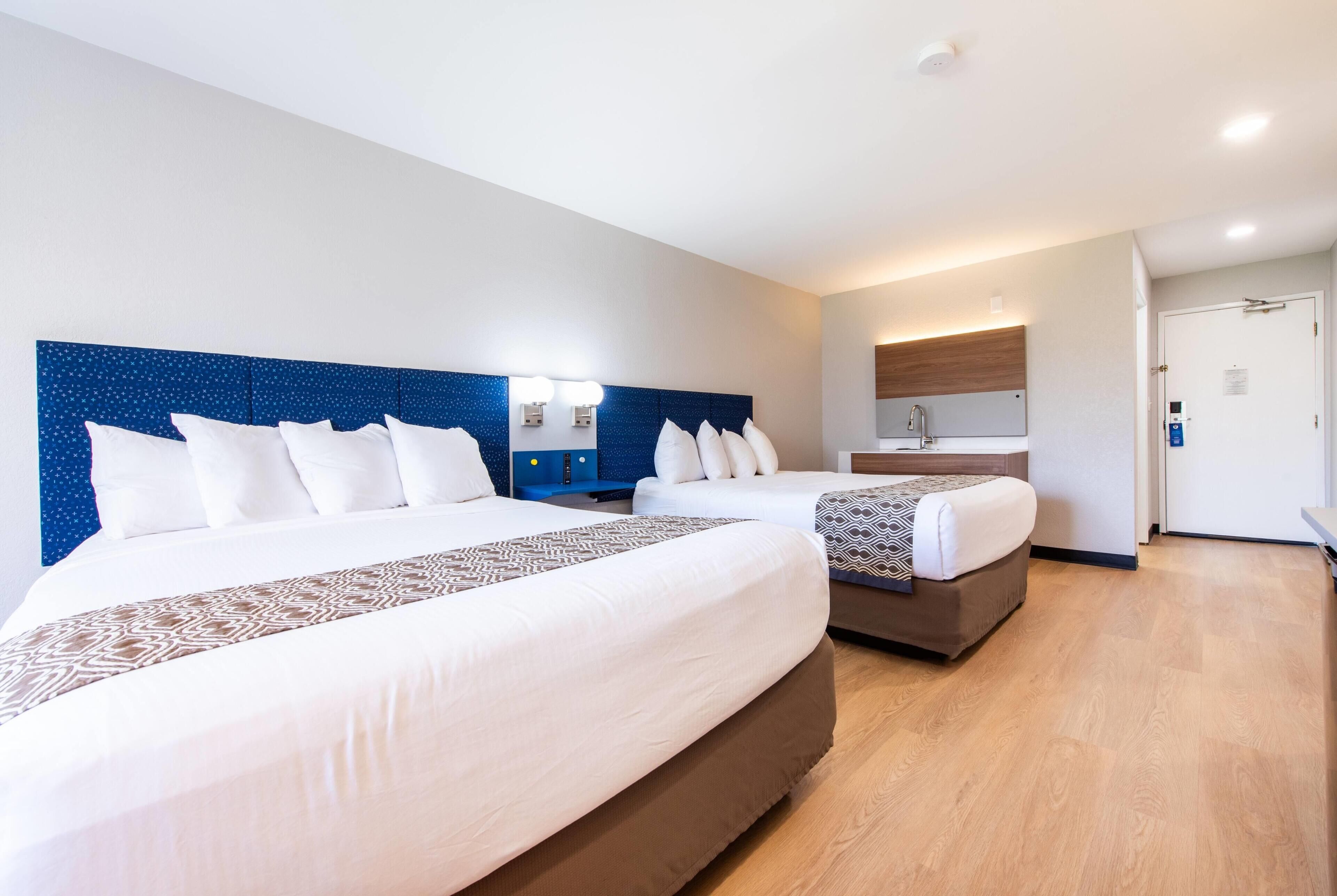 Habitación de hotel con dos camas, kitchenette pequeña, baño y puerta.