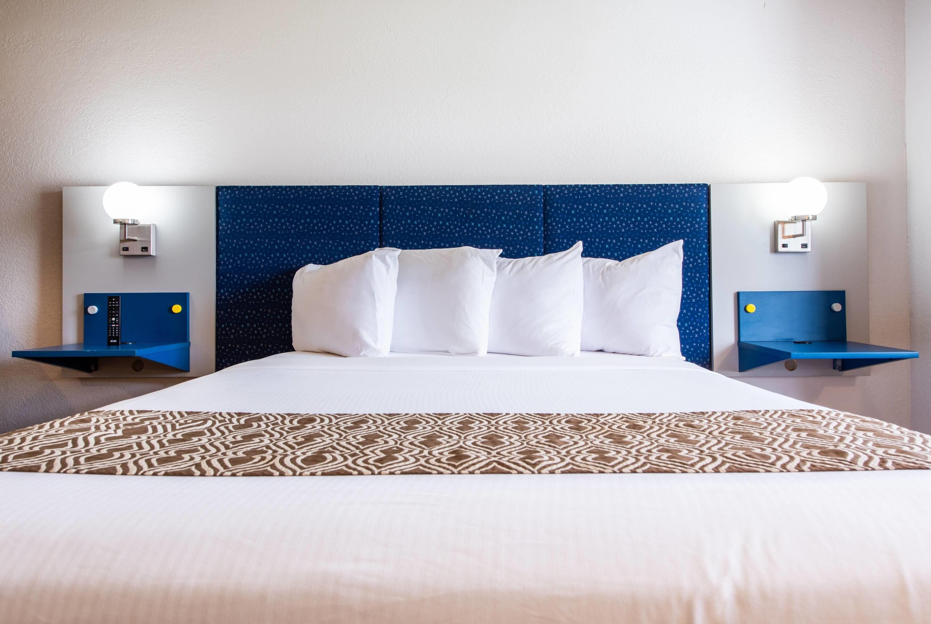 A hotel room with a large bed, white linens, a patterned throw, a blue headboard, and wall-mounted nightstands with lights.