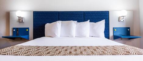 A hotel room with a large bed, white linens, a patterned throw, a blue headboard, and wall-mounted nightstands with lights.
