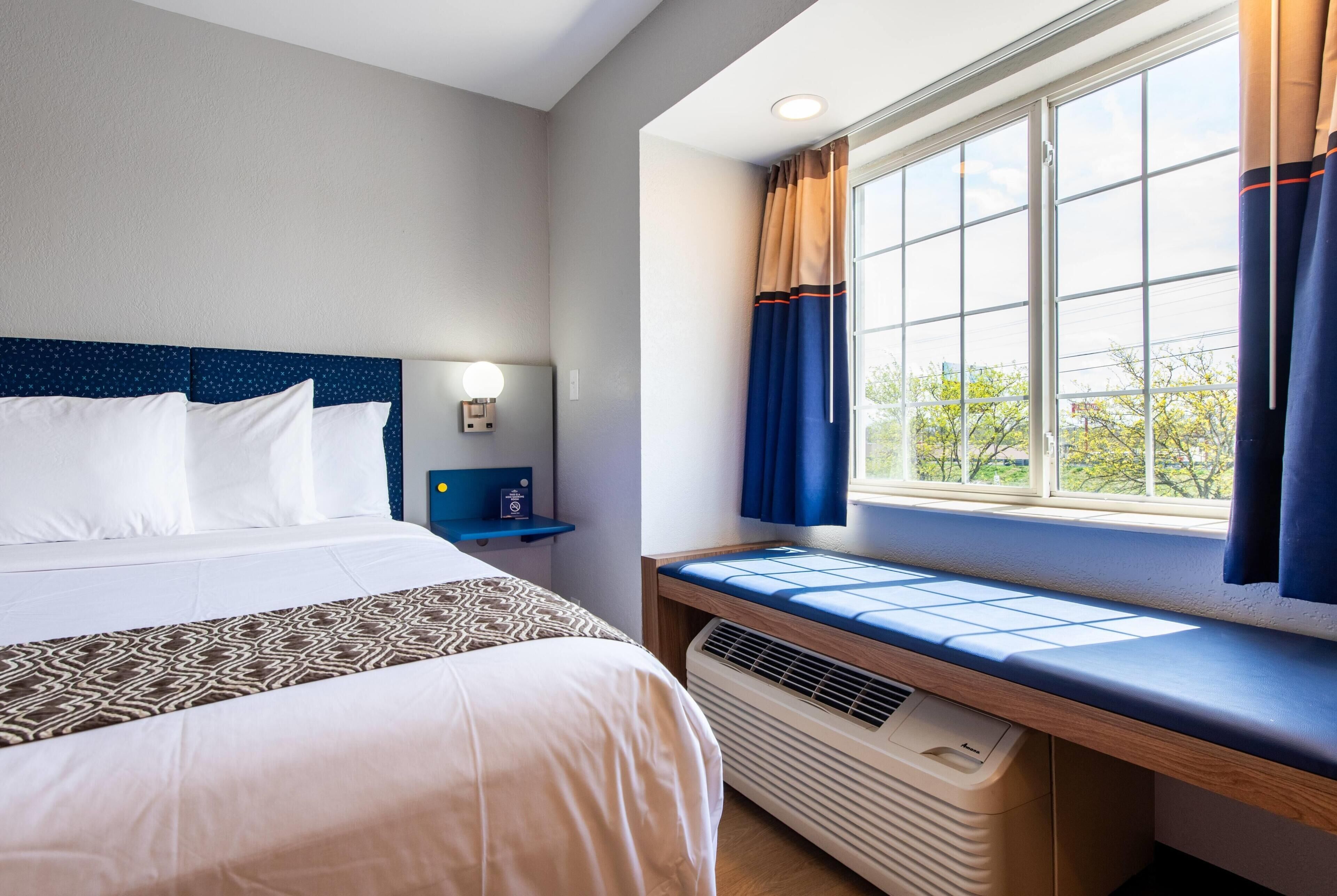 A hotel room with a bed, bedside table, lamp, window with curtains, and a view of trees.