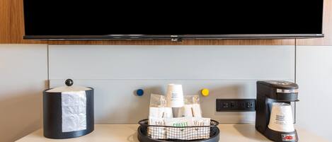 A hotel room with a flat-screen TV mounted on the wall, a coffee maker, and a basket of coffee packets on the counter.
