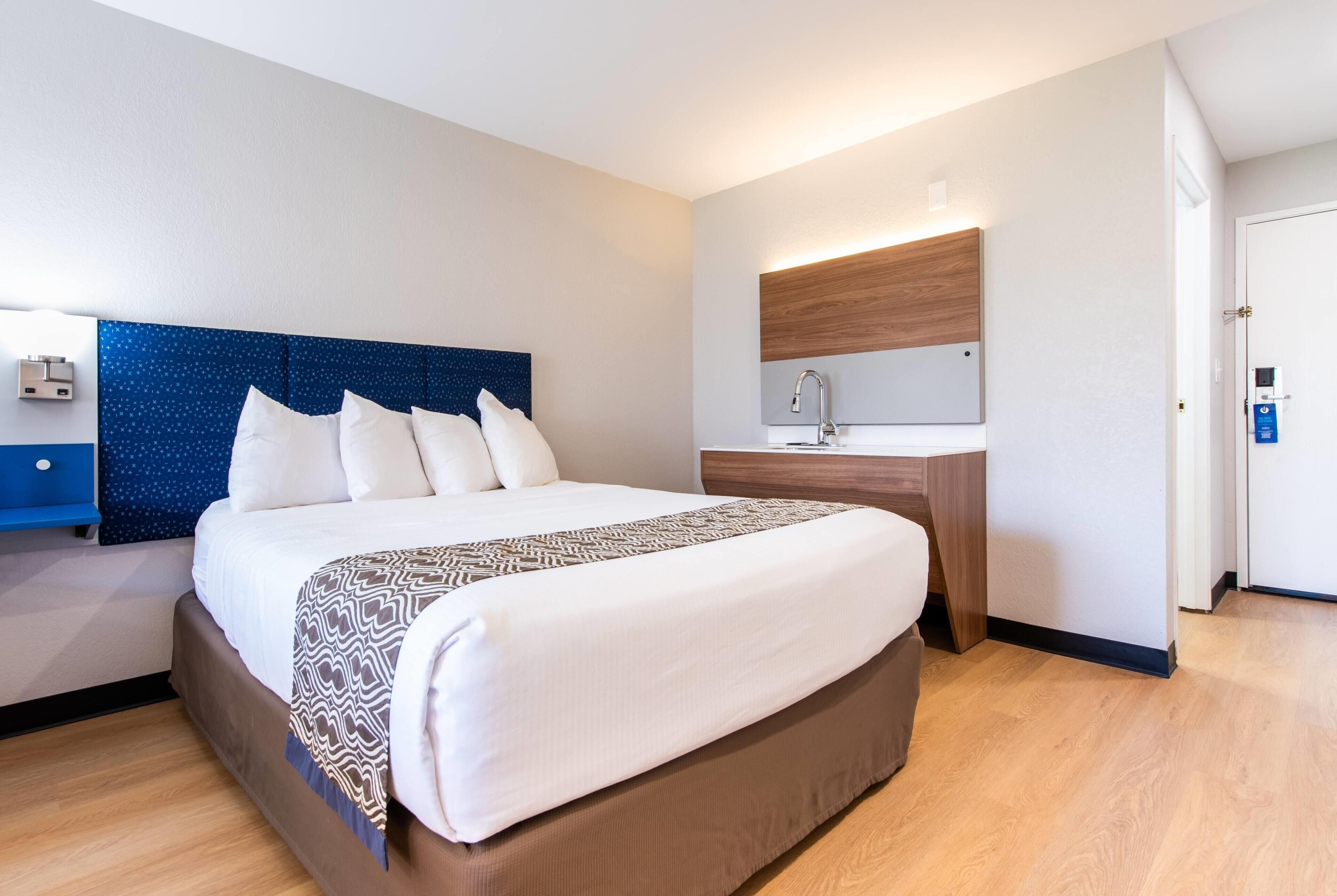 A hotel room with a large bed, a bedside table, a built-in desk, a mirror, and a door.