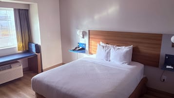 A hotel room with a large bed, a desk with a chair, a window with curtains, a wall-mounted lamp, and a ceiling fan.