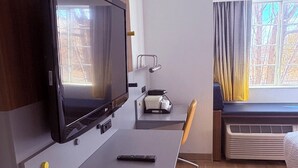 A hotel room with a flat-screen TV mounted on a wall, a desk with a remote control, a small table with a tissue box, a chair, and a window with curtains.