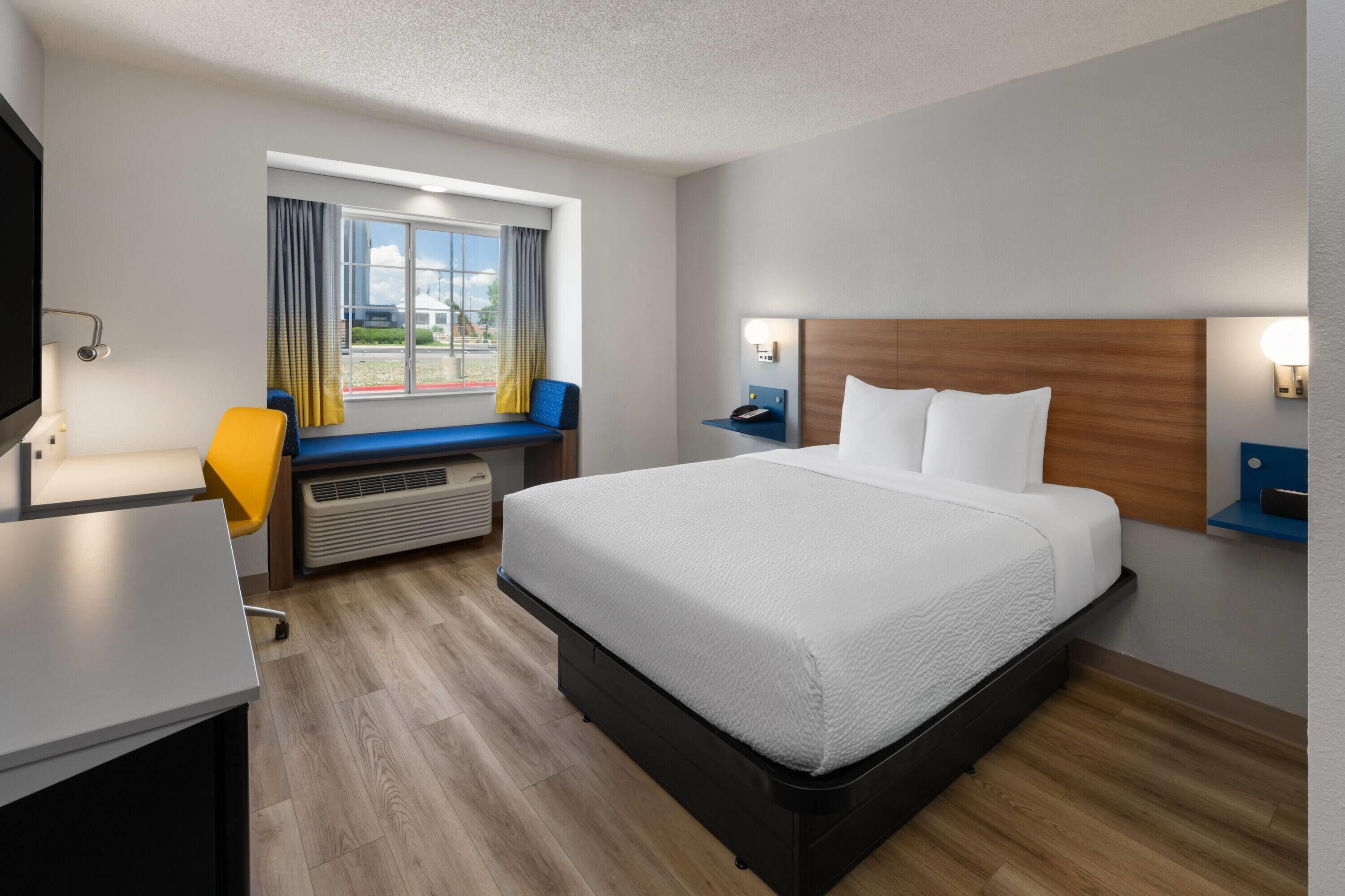 Una habitación de hotel con cama, escritorio, silla, ventana con cortinas y una lámpara fijada a la pared.