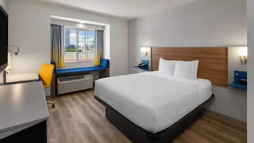 Una habitación de hotel con cama, escritorio, silla, ventana con cortinas y una lámpara fijada a la pared.