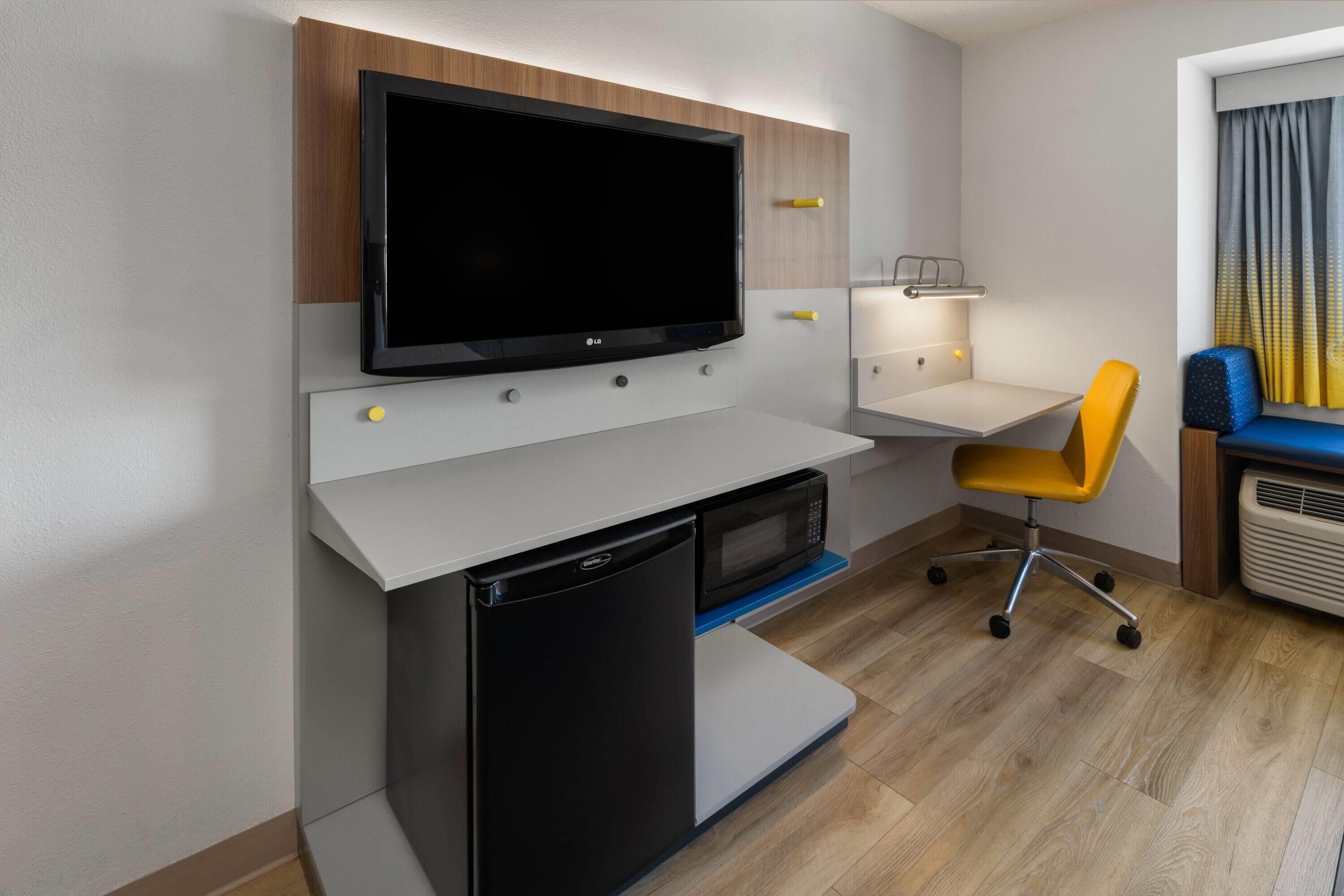 A compact hotel room with a flat-screen TV mounted on a wall, a small refrigerator, a desk with a chair, and a window with curtains.
