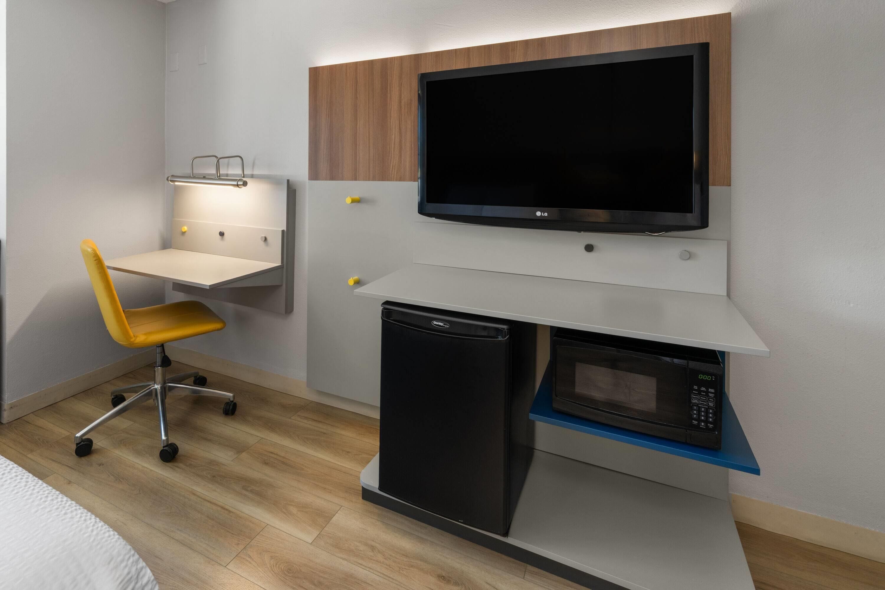 A compact hotel room with a bed, a desk with a chair, a television, a microwave, and a dishwasher.