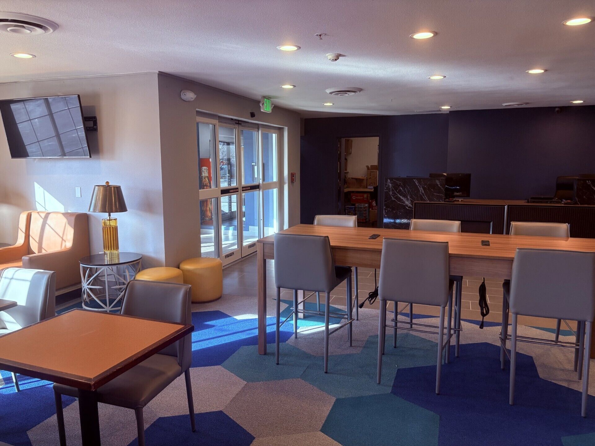 A modern hotel lobby with a large wooden table, comfortable seating, a television, and a glass door leading to a balcony.