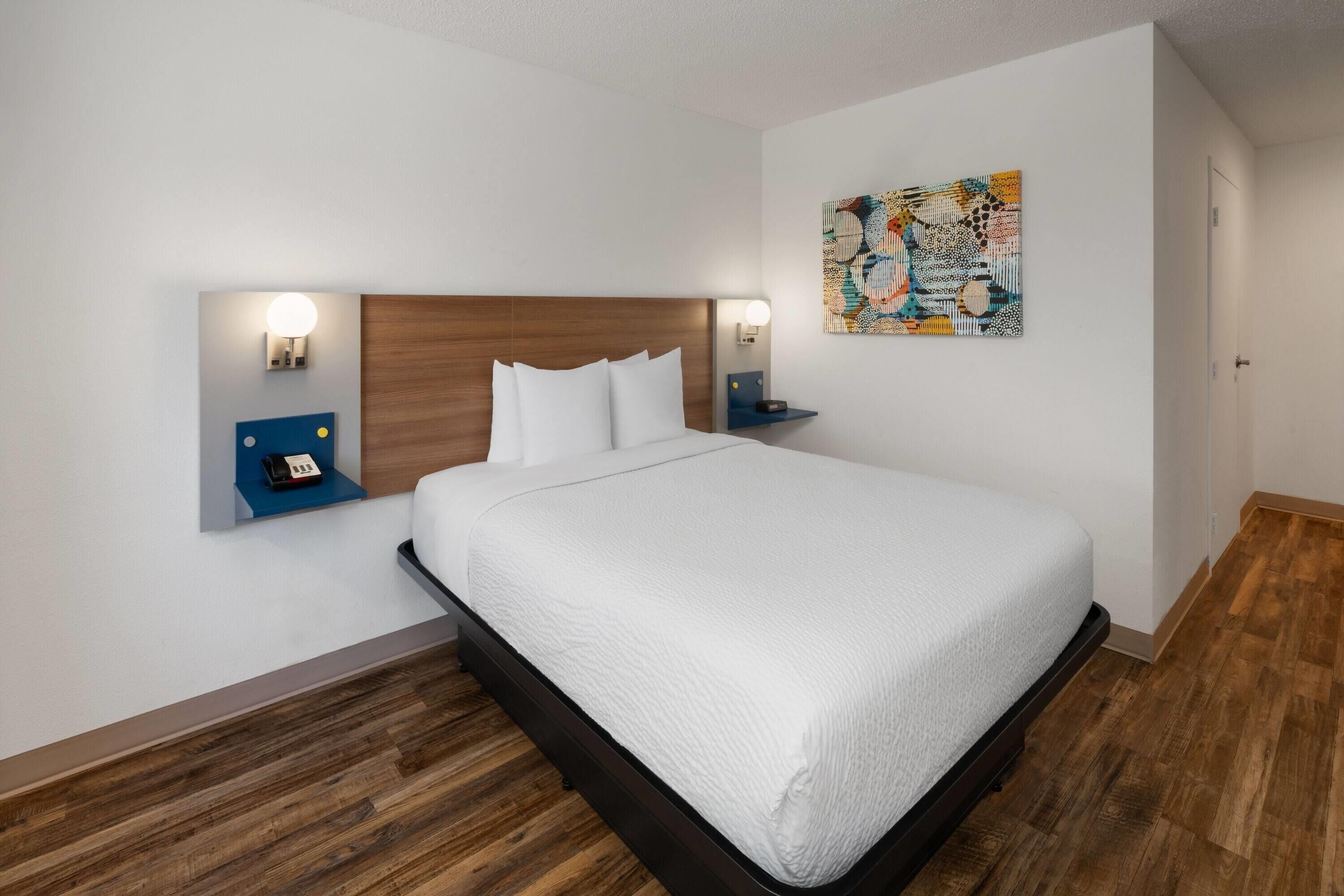 A hotel room with a large bed, wooden headboard, two bedside tables with lamps, a wall-mounted phone, and a colorful abstract painting.