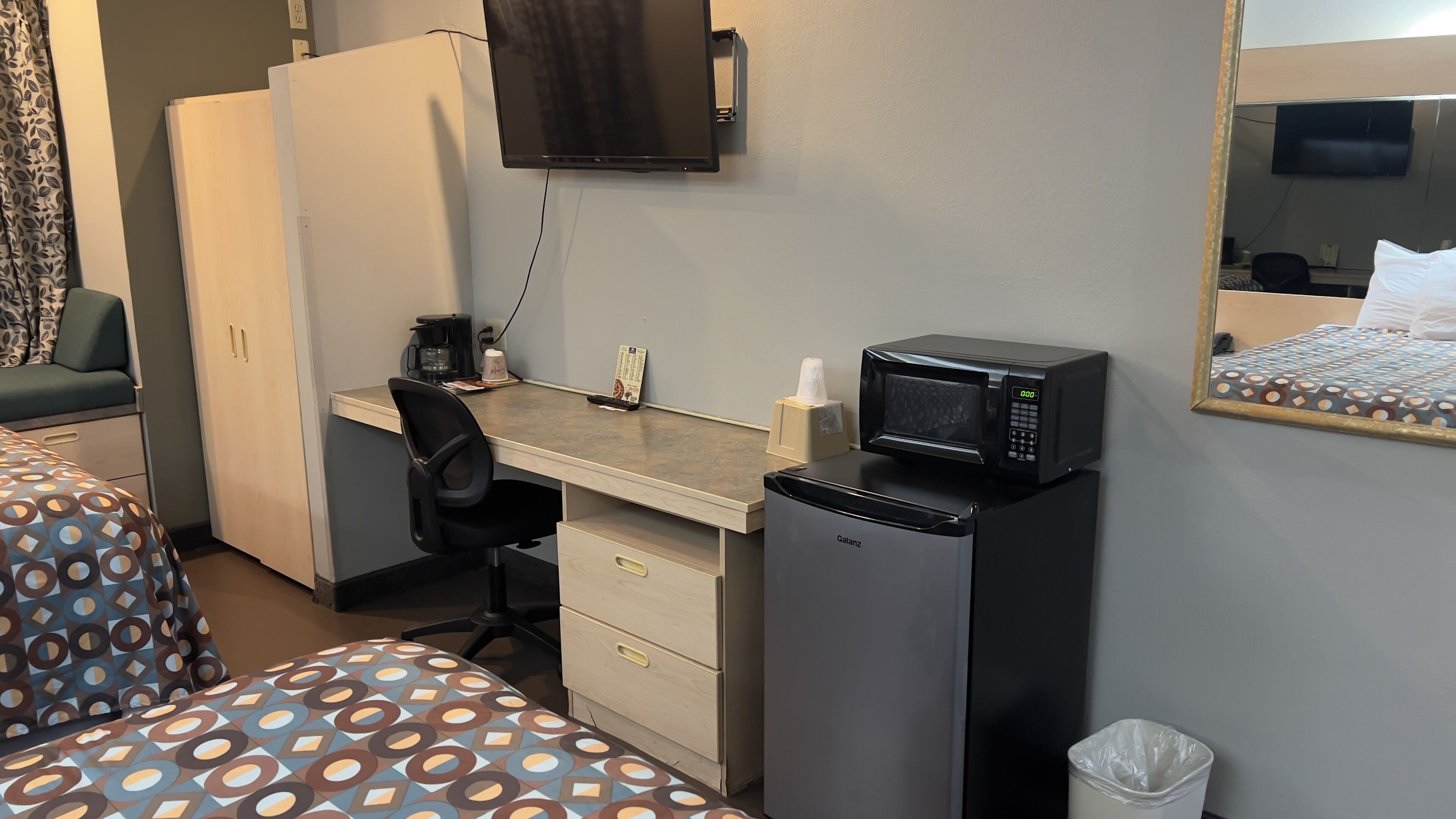 A hotel room with a desk, a chair, a bed, a TV, a microwave, and a trash bin.