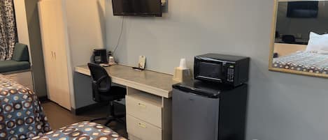A hotel room with a desk, a chair, a bed, a TV, a microwave, and a trash bin.