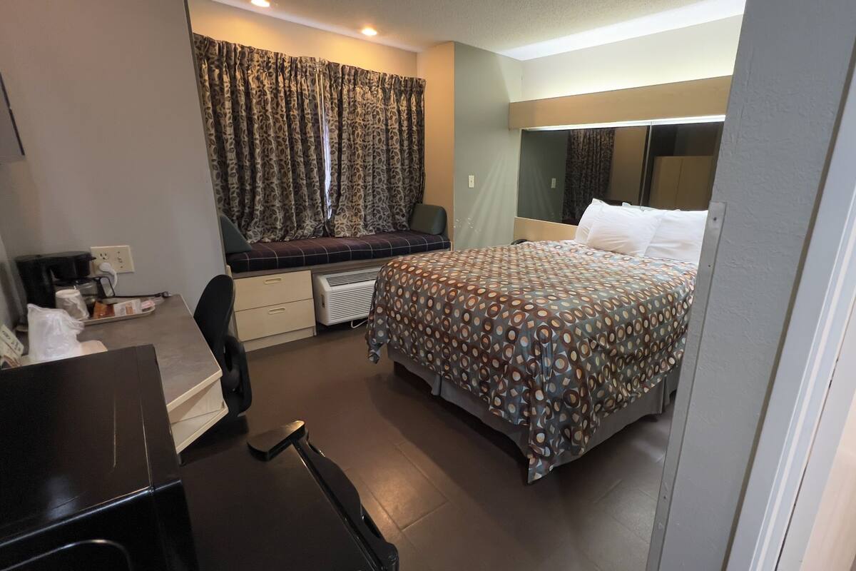 Ein Hotelzimmer mit Bett, Schreibtisch mit Computer, Stuhl, Fenster mit Vorhängen und Nachttisch.